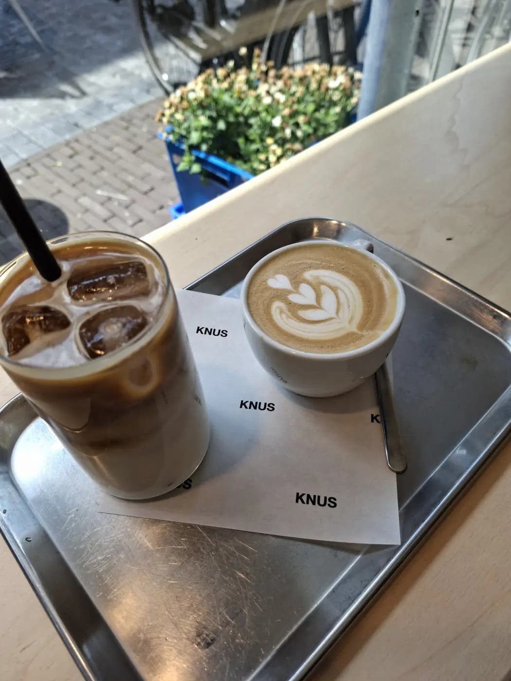 Iced latte en cappuccino op een stalen dienblad aan het raam, met de straat erachter.